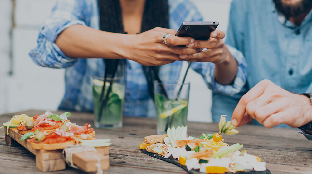A couple eating a meal with one of them taking a photo of their plate using their phone