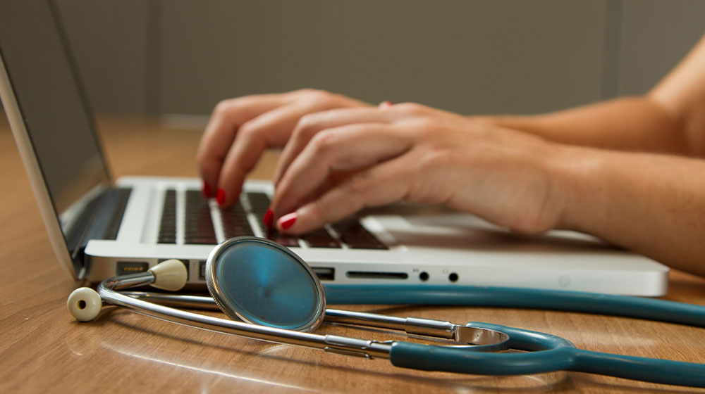 Close up of hands typing on a laptop next to a stethescope