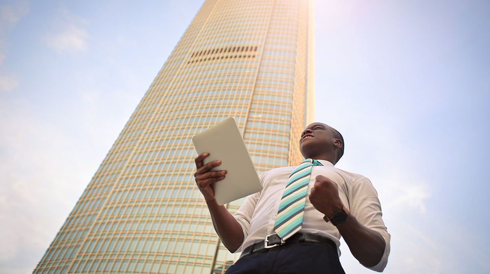 A man in a shirt and tie stands outside a tall skyscraper while holding a tablet and gesturing enthusiastically A man in a shirt and tie stands outside a tall skyscraper while holding a tablet and gesturing enthusiastically