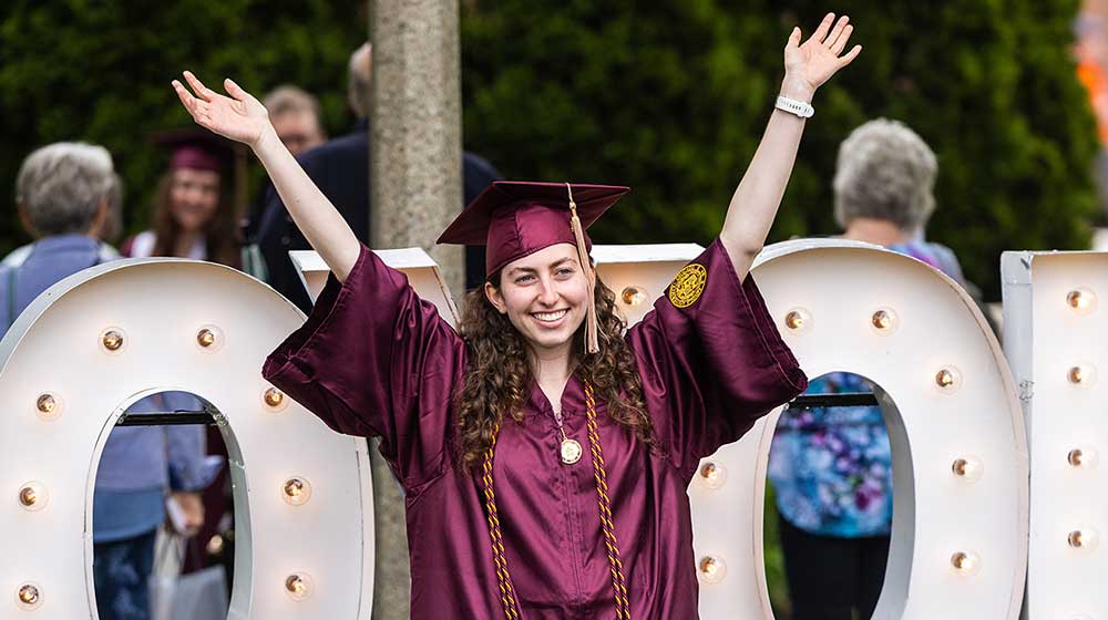 A student in maroon graduation robes lifting her arms in joy