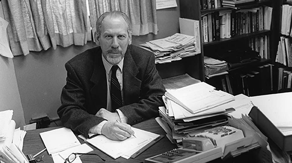 An archival photo of George Kaufman sitting in his office surrounded by books and papers An archival photo of George Kaufman sitting in his office surrounded by books and papers