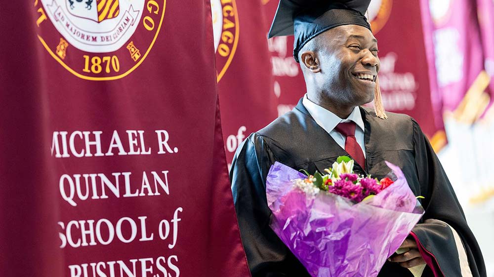 Person in graduation hat and gown smiling and holding flowers next to a maroon banner that says 