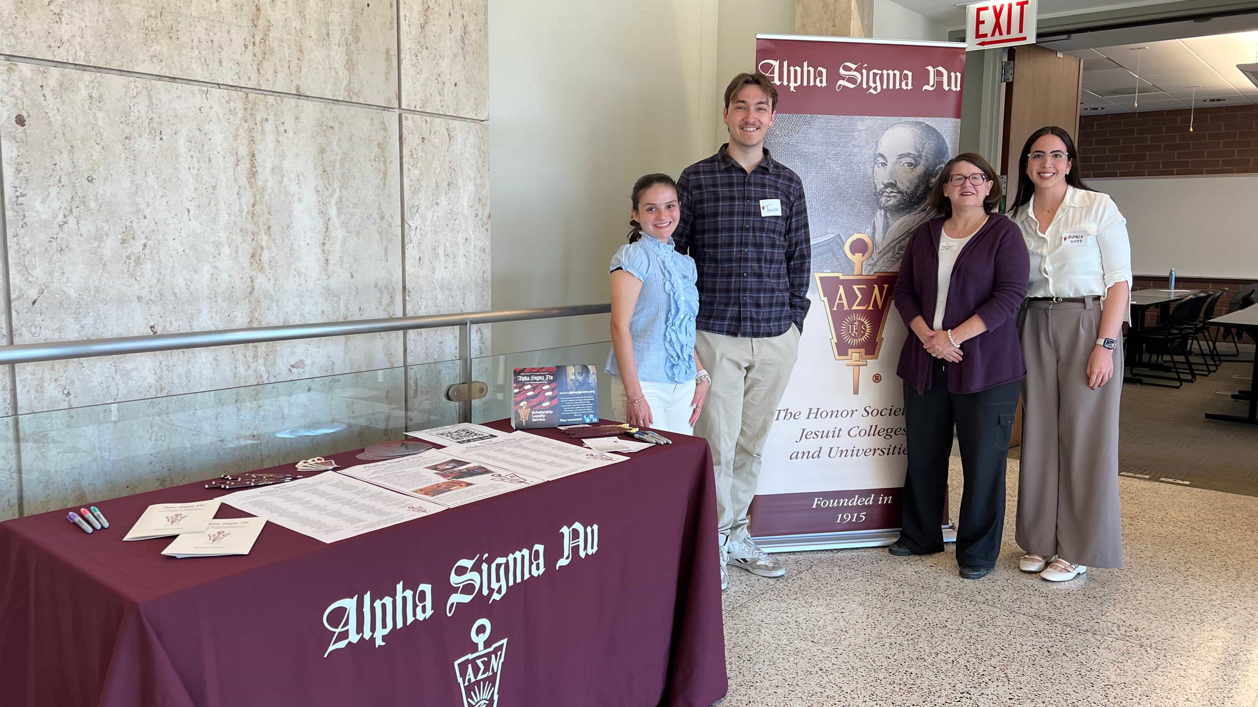 Two student leaders and two alumni members of Alpha Sigma Nu stand in front of the ASN banner and table with ASN materials.