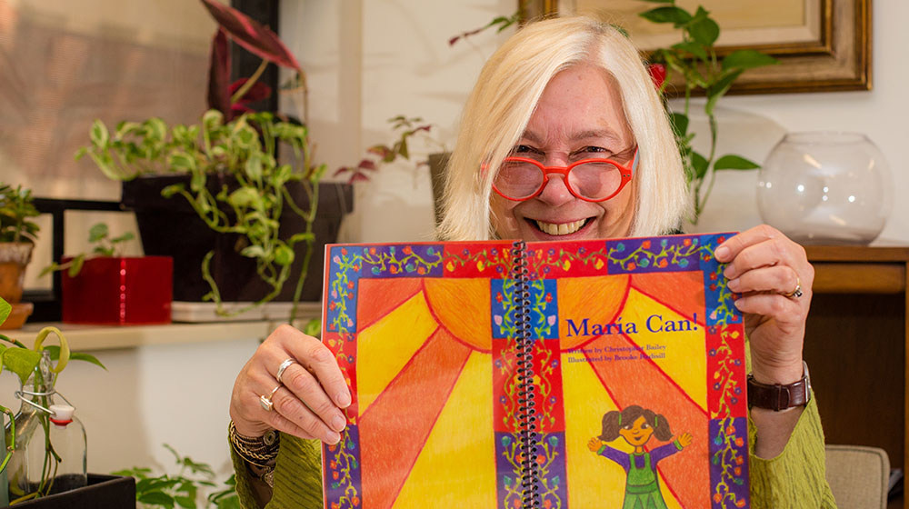 Loyola professor Bren Murphy, PhD, poses with one of the children’s books that her students created in her Community as Story class. “We’re trying to use this class to look at the power of storytelling in communities to say what’s normal and what isn’t—because that’s what books do,” she said. (Photo: Natalie Battaglia)

