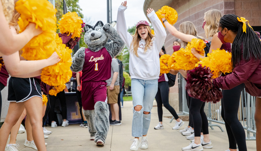  A group of cheerleaders with yellow pom-poms forms a tunnel as a person in a wolf mascot costume and a young woman in casual clothes run through, celebrating.