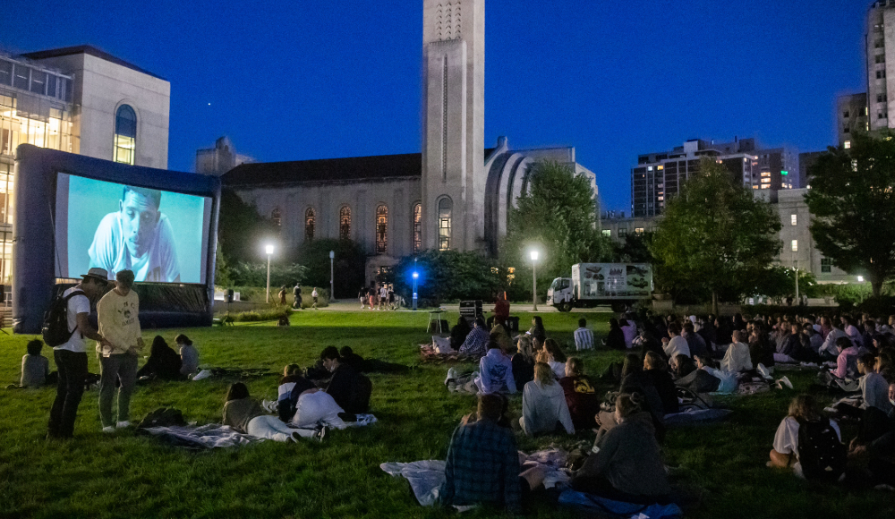  A large group of people sitting on a grassy field at night, watching a movie projected onto a screen.