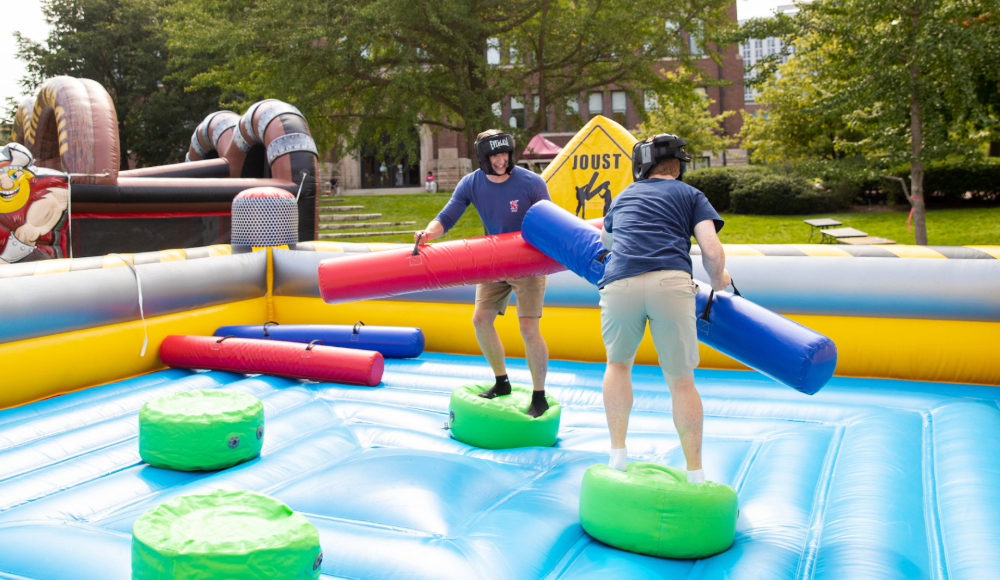 Two people wearing helmets balance on pedestals in an inflatable jousting arena, each holding a padded joust stick, outdoors on a sunny day.
