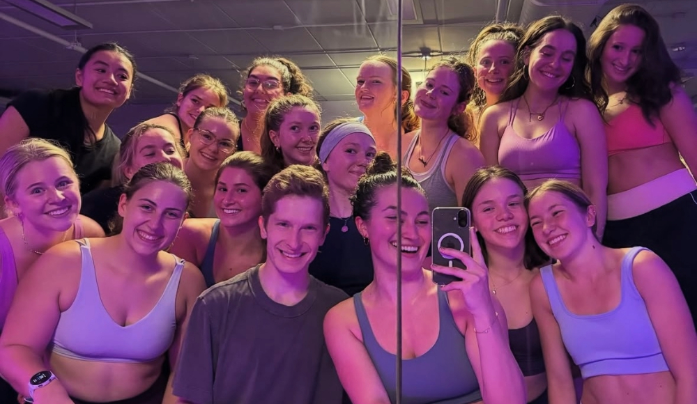 A diverse group of young adults poses for a selfie in a brightly lit studio with purple and pink lighting.
