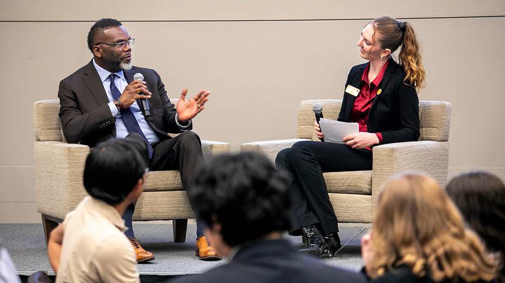 Erin Tylutki speaks with Mayor Brandon Johnson from armchairs on an elevated stage as students spectate.