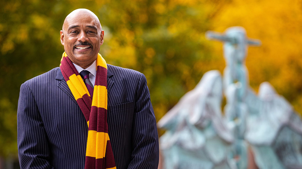 Keith M. Champagne stands on campus during a fall day, with foliage and the wolf and kettle statue.