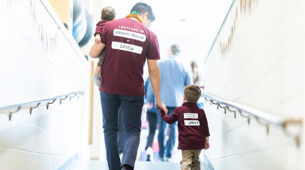 Brian Tabata wearing a maroon t-shirt stating where he matched for residency carrying one of his sons and holding the hand of his other son, both wearing maroon shirts to match their dad. Brian Tabata wearing a maroon t-shirt stating where he matched for residency carrying one of his sons and holding the hand of his other son, both wearing maroon shirts to match their dad.